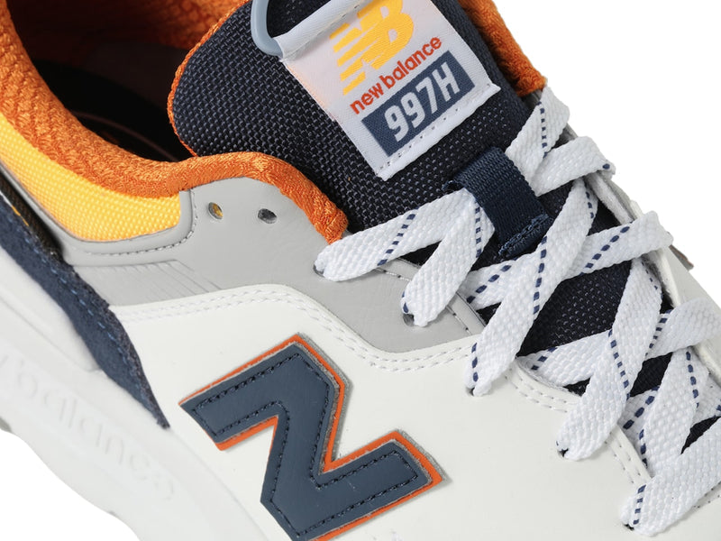 New Balance 997H sneakers with white leather upper, navy and orange accents, and white braided laces, close-up view.