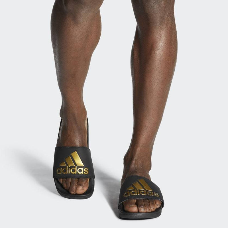 Adidas Adilette Comfort Slides in black with gold logo branding and cushioned footbed, front view worn by individual.