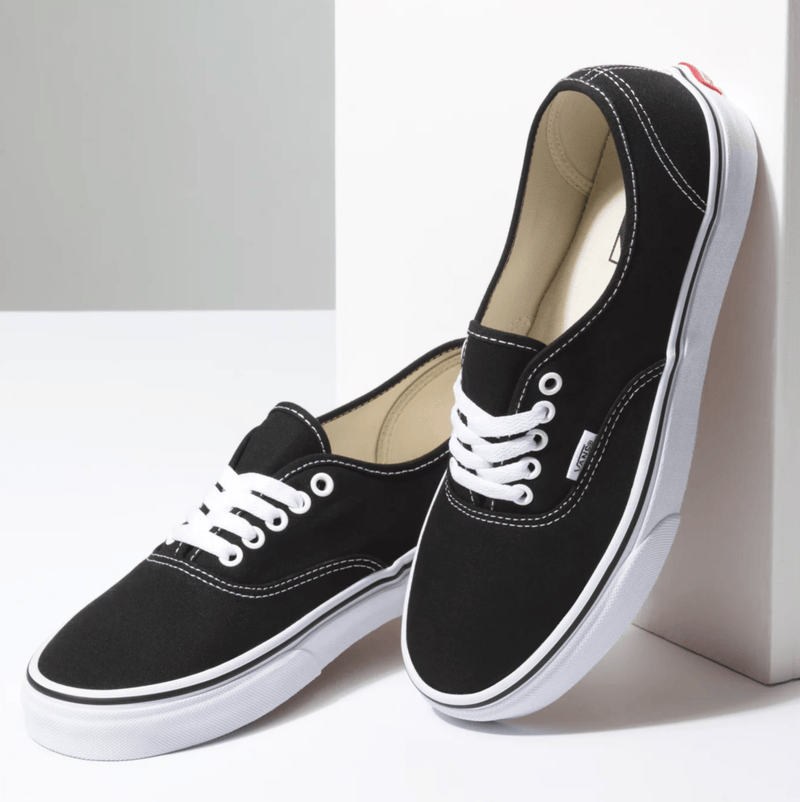 Vans Authentic sneakers in black canvas with white stitching, white laces, and vulcanized rubber outsole, angled side view.
