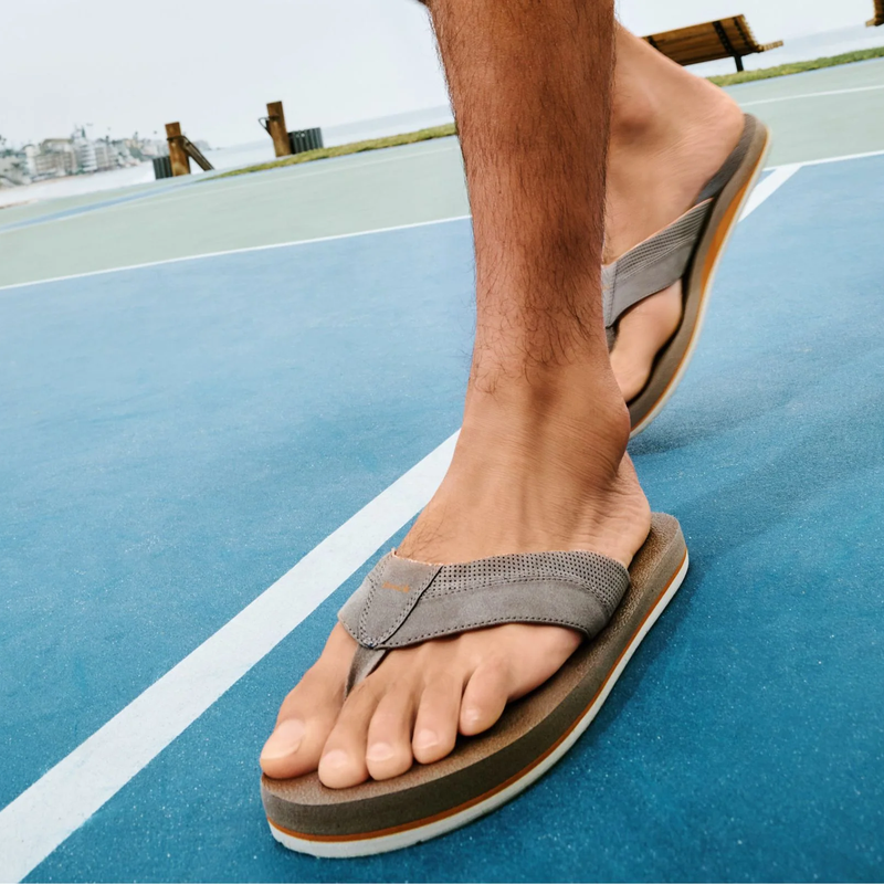 Sanuk Beer Cozy IV sandals in gray with perforated straps and cushioned footbed, worn outdoors on a blue court.