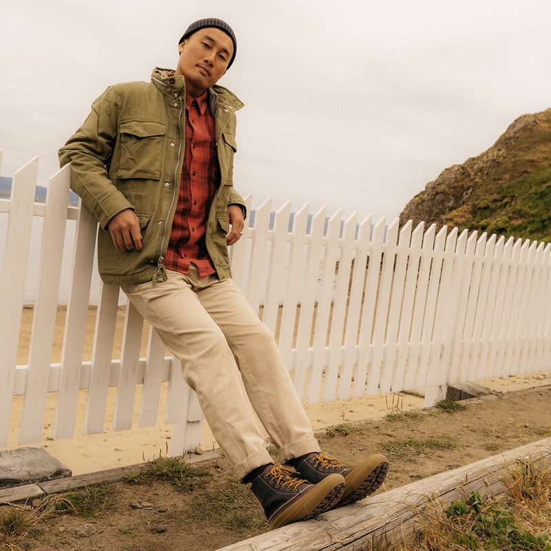 SeaVees Cascade Range Boot in black leather with brown laces and lugged outsole, outdoor lifestyle setting with wooden and sandy terrain.