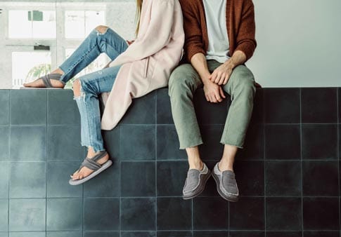 Sanuk Chiba casual slip-on shoes in gray with textured canvas upper and visible stitching detail, lifestyle setting with seated individuals.