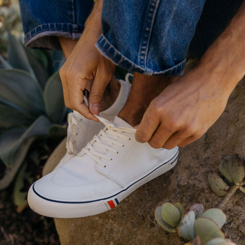 SeaVees Coastal Cruiser sneakers in white canvas with blue and red detailing on the sole, lifestyle view of person lacing shoes.