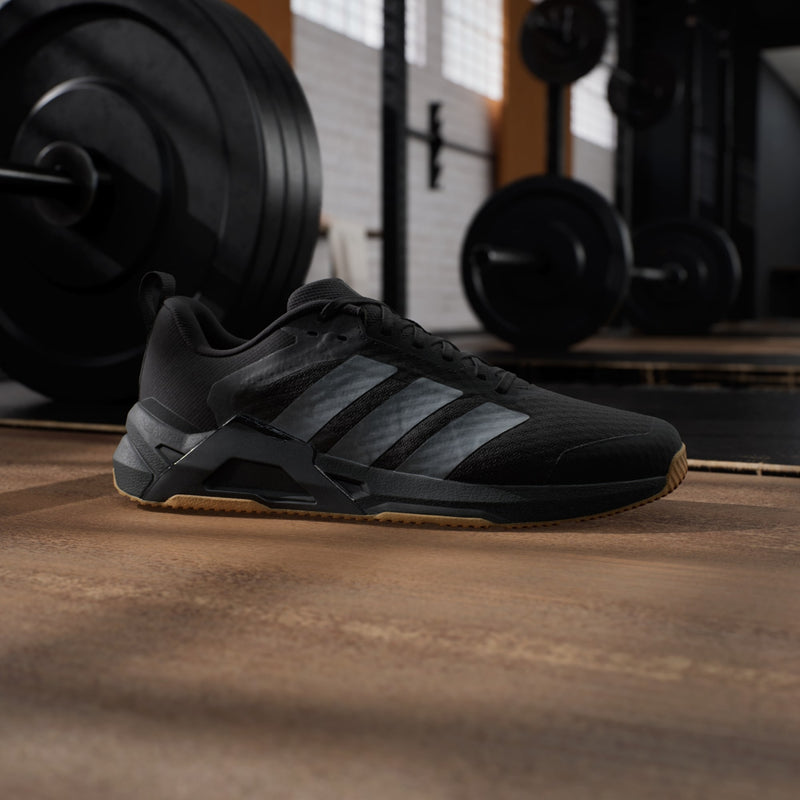 Adidas Dropset Control Trainer shoes in black with gray stripes and gum sole, side view in gym setting.