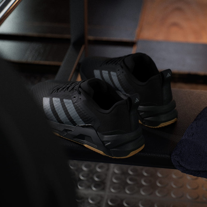 Adidas Dropset Control Trainer shoes in black with gray stripes and gum sole, gym setting rear perspective.