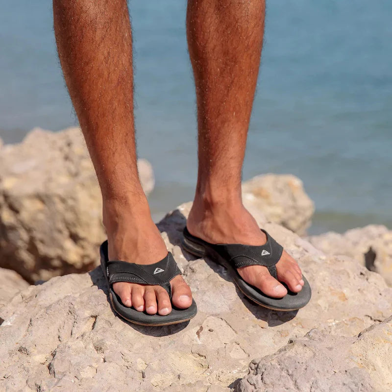 Reef Fanning Flip Flops in black with padded straps and signature arch support, worn outdoors on rocky terrain by the ocean.