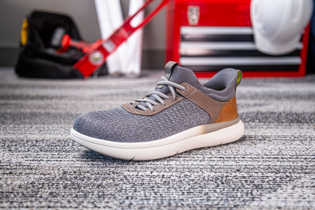 Florsheim Work Fleet Work Comp Toe Sneaker in gray with safety toe, brown heel accents, and cushioned white sole, side view on work setting carpet.