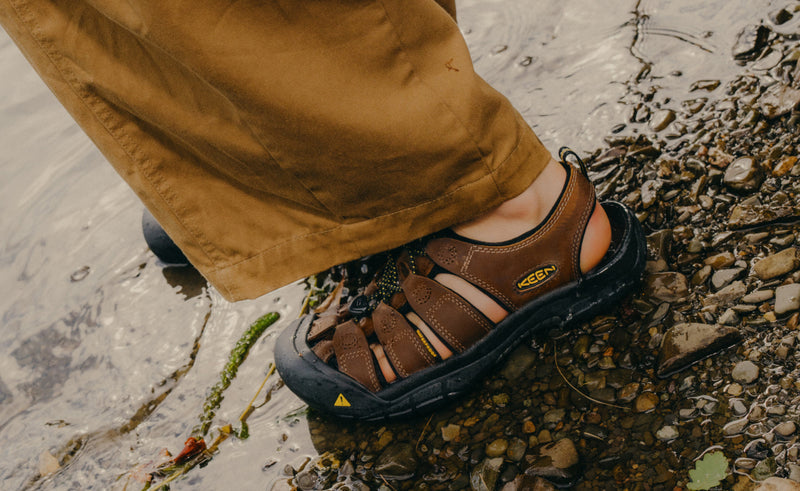 Keen Newport Leather sandals in brown leather with black sole and breathable cutouts, worn outdoors by water on rocky terrain.