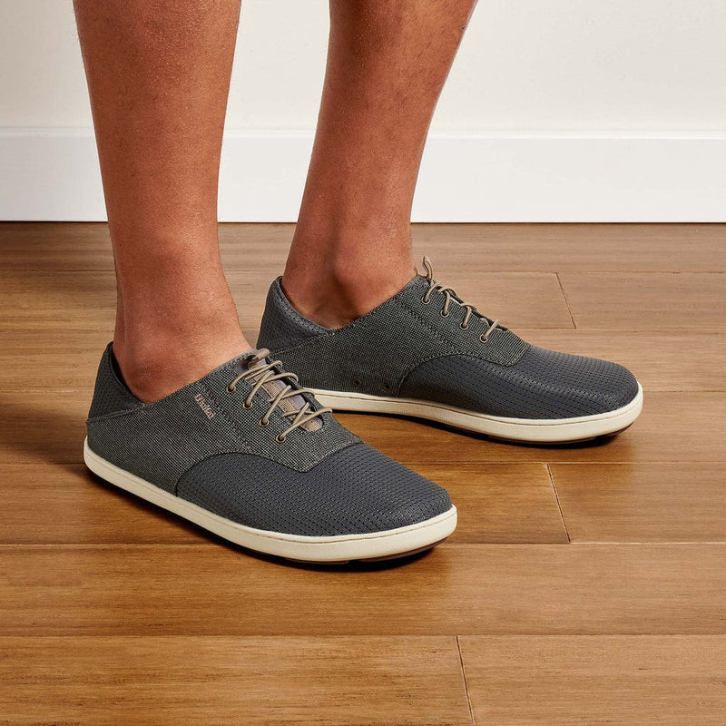 OluKai Nohea Moku casual shoes in dark gray with lightweight mesh upper, contrast white sole, and lace-up design, worn by a person indoors.