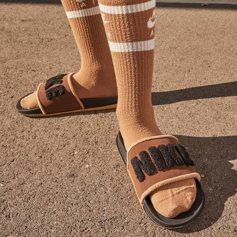 Nike Offcourt Slide SE sandals in brown with plush "NIKE" text detail on strap, worn outdoors with socks.
