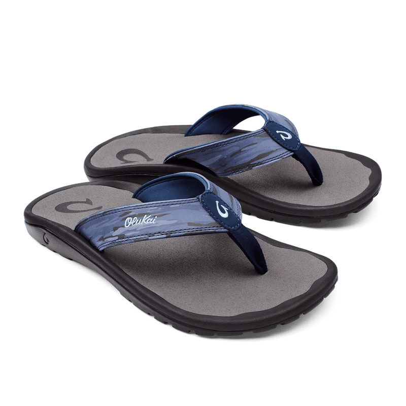 OluKai Ohana Pa'i sandals with blue and gray water-inspired printed straps, cushioned footbed, and black rubber sole, angled view.