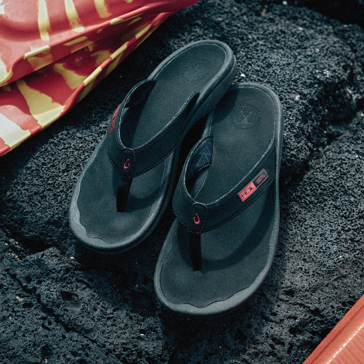 OluKai ‘Ohana sandals in black with red logo accents and water-resistant synthetic straps, top view on rocky surface.