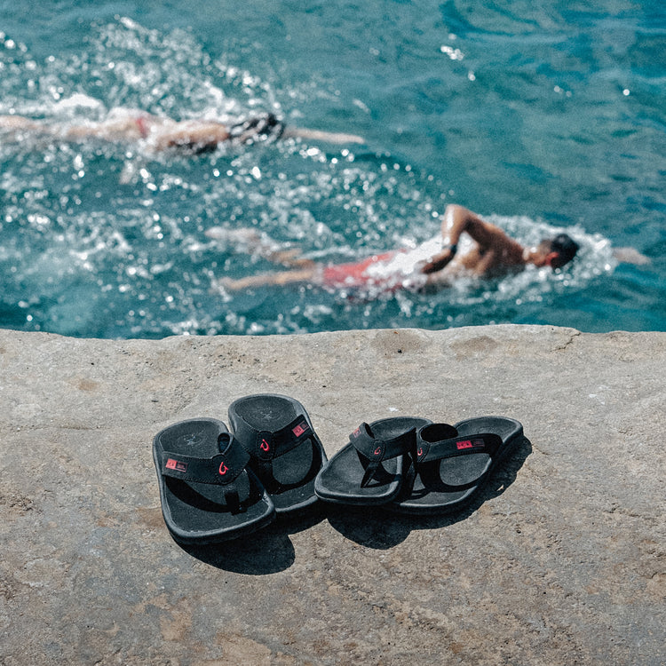 OluKai ‘Ohana sandals in black with red logo accents and water-resistant straps, pictured on stone near swimmers in the ocean.
