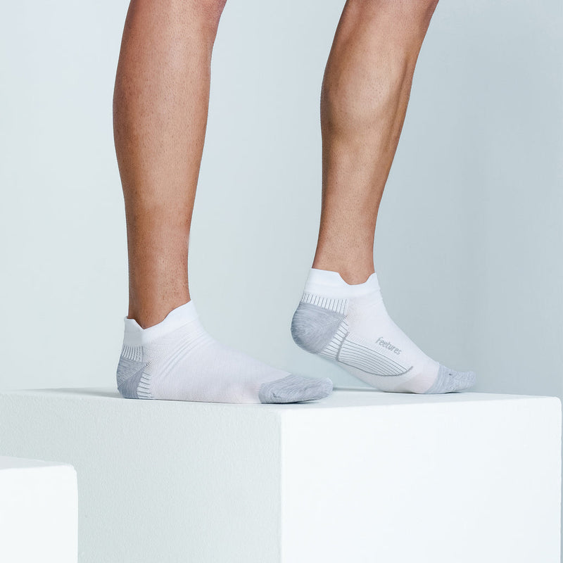 Feetures Plantar Fasciitis Relief Sock Light Cushion No Show Tab in white and gray with targeted compression, worn by a person standing on a platform.