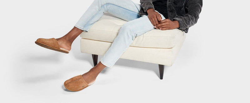 UGG Scuff slippers in chestnut suede with wool lining, worn by seated person on a beige ottoman.