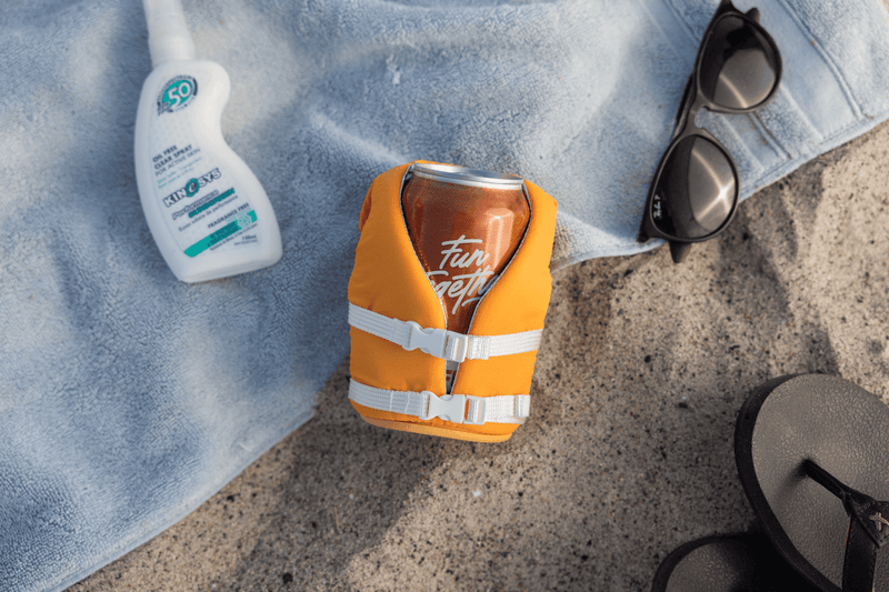 Puffin The Buoy drink holder in orange designed like a life vest with white straps and buckles, beach lifestyle setting.