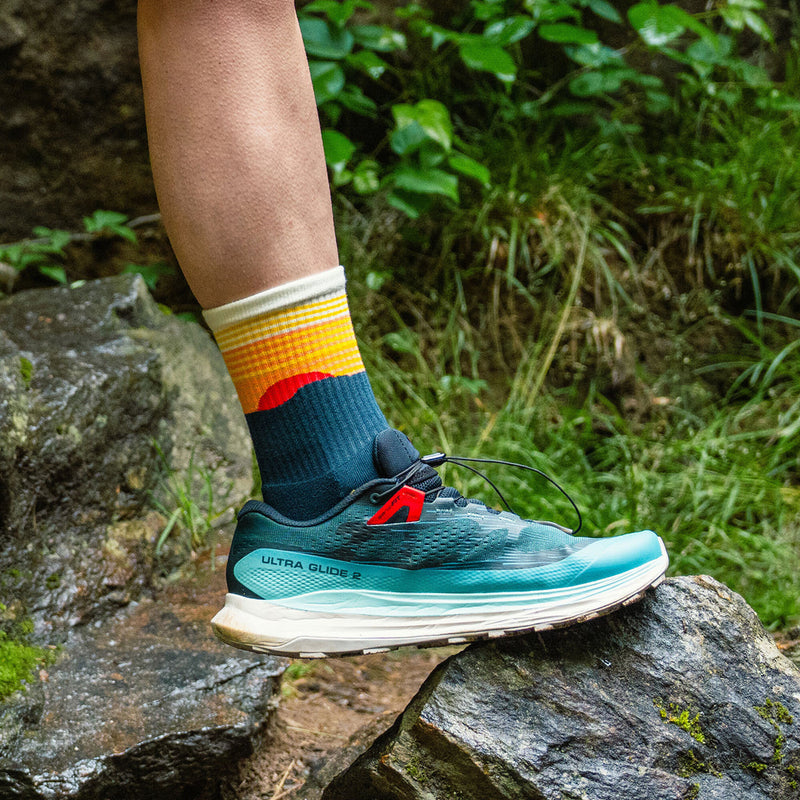 Feetures Trail Max Cushion Mini Crew Sock in navy with yellow and orange gradient design, worn by hiker outdoors on rocky terrain.