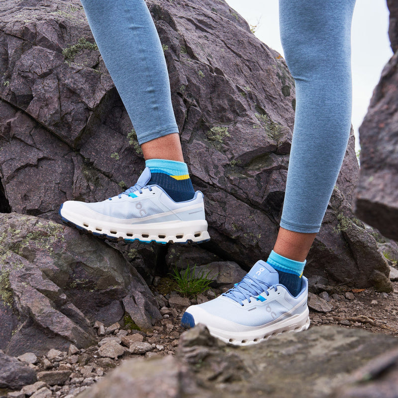 "Feetures Trail Max Cushion Quarter Sock in navy blue with yellow and light blue accents, worn by hiker on rocky terrain"