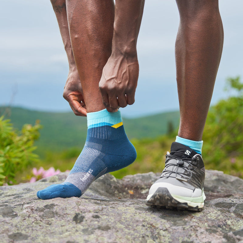 Feetures Trail Max Cushion Quarter Sock in blue and aqua with yellow accents and durable cushioning, worn by hiker outdoors.